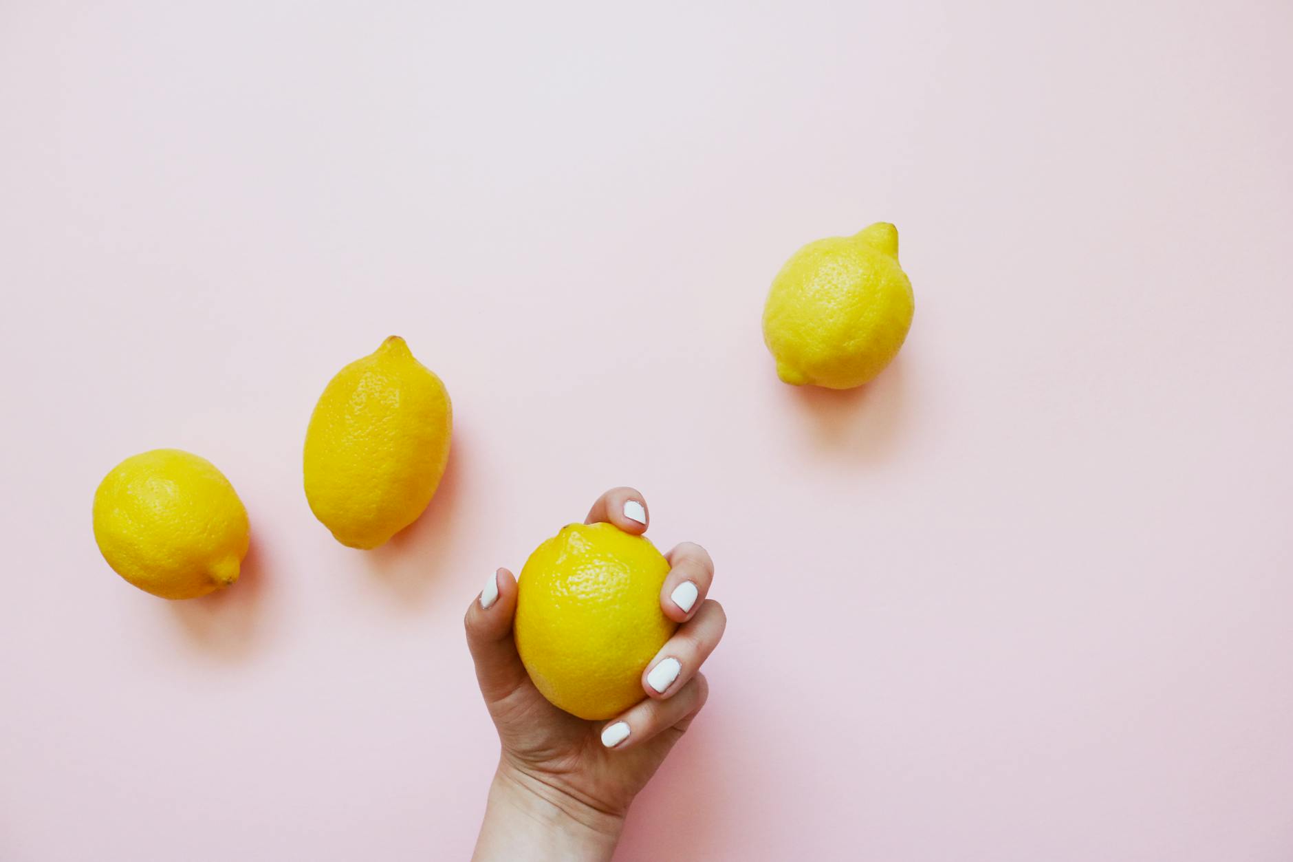 Hand holding a bright lemon on soft pink background with yellow lemons arranged nearby