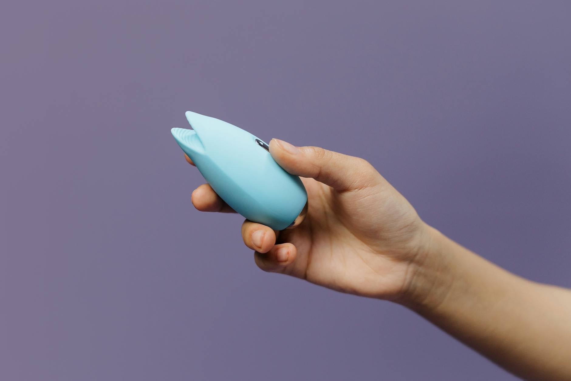 Hand holding a blue silicone clitoral vibrator against a purple background, representing pleasure and self-care.