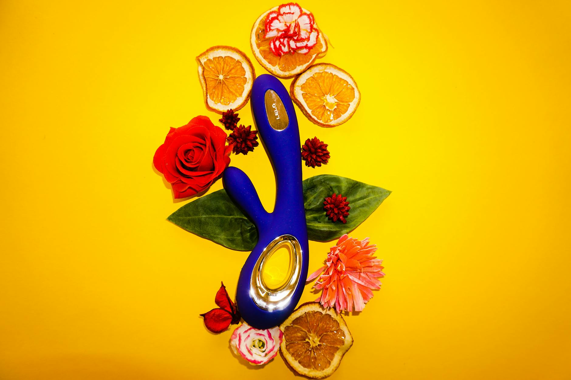 A blue vibrator surrounded by flowers and citrus on a yellow background, symbolizing sensual rediscovery